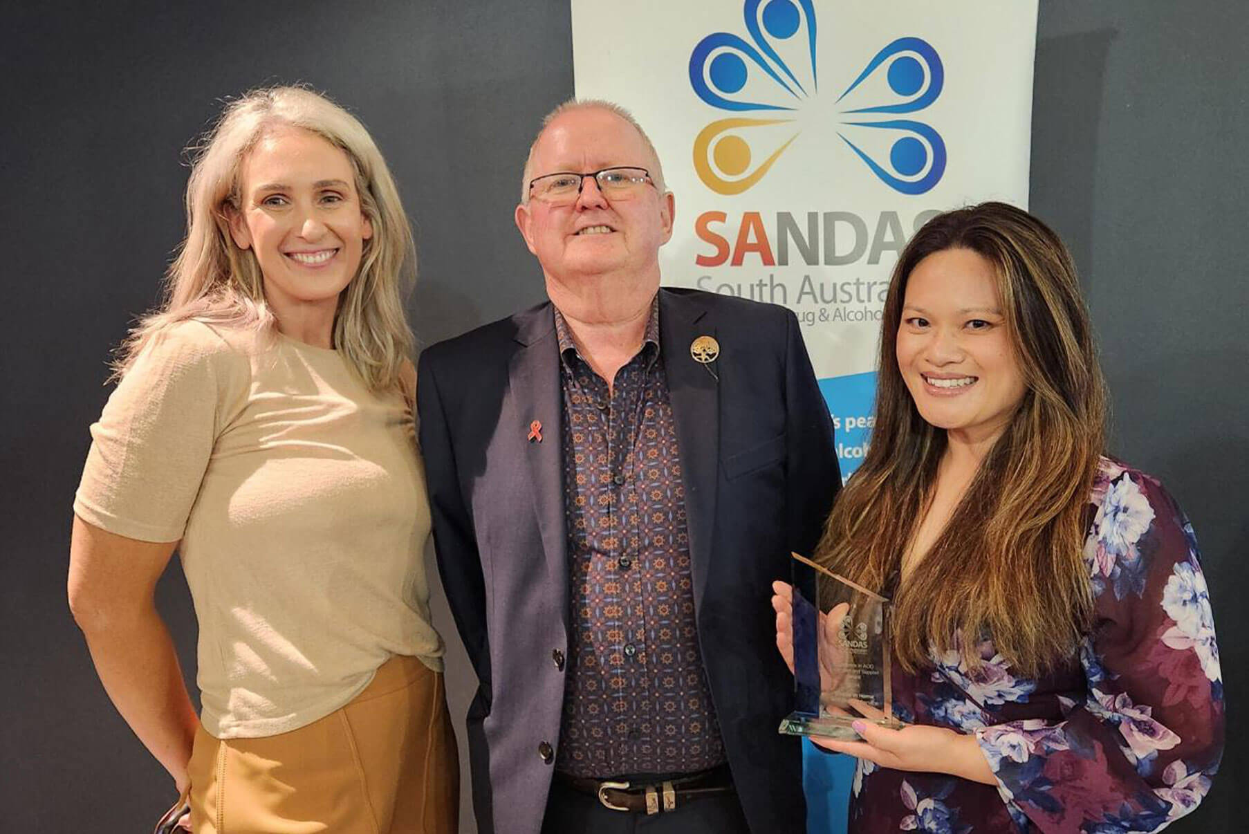 Three people stand together, with one woman holding a clear award plaque in front of a branded banner.