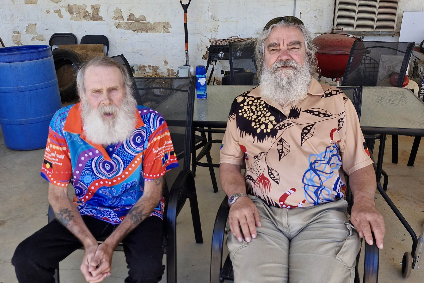 Two elderly, bearded men sit together in chairs outdoors on a sunny day.