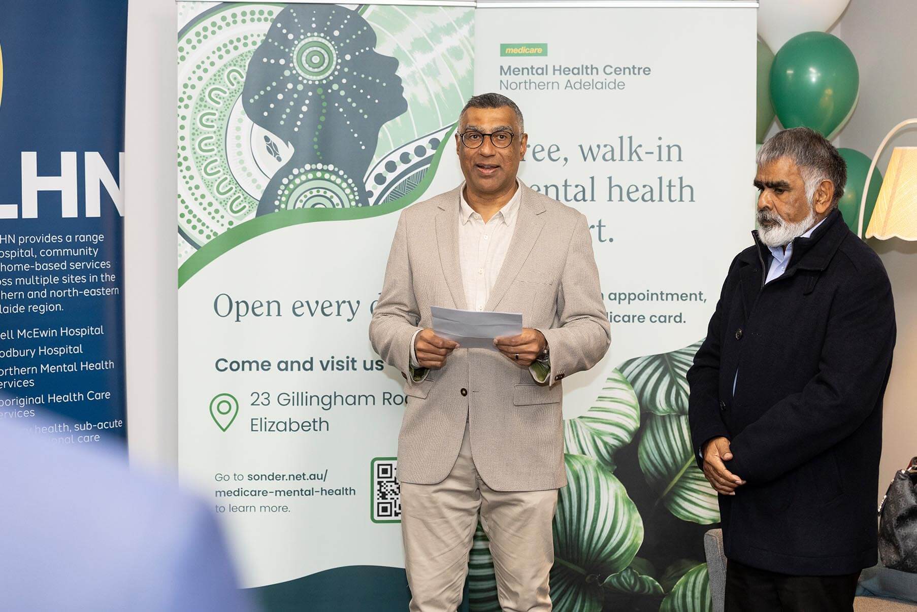 A man in a suit jacket holds a paper while standing next to another man in front of an event banner.