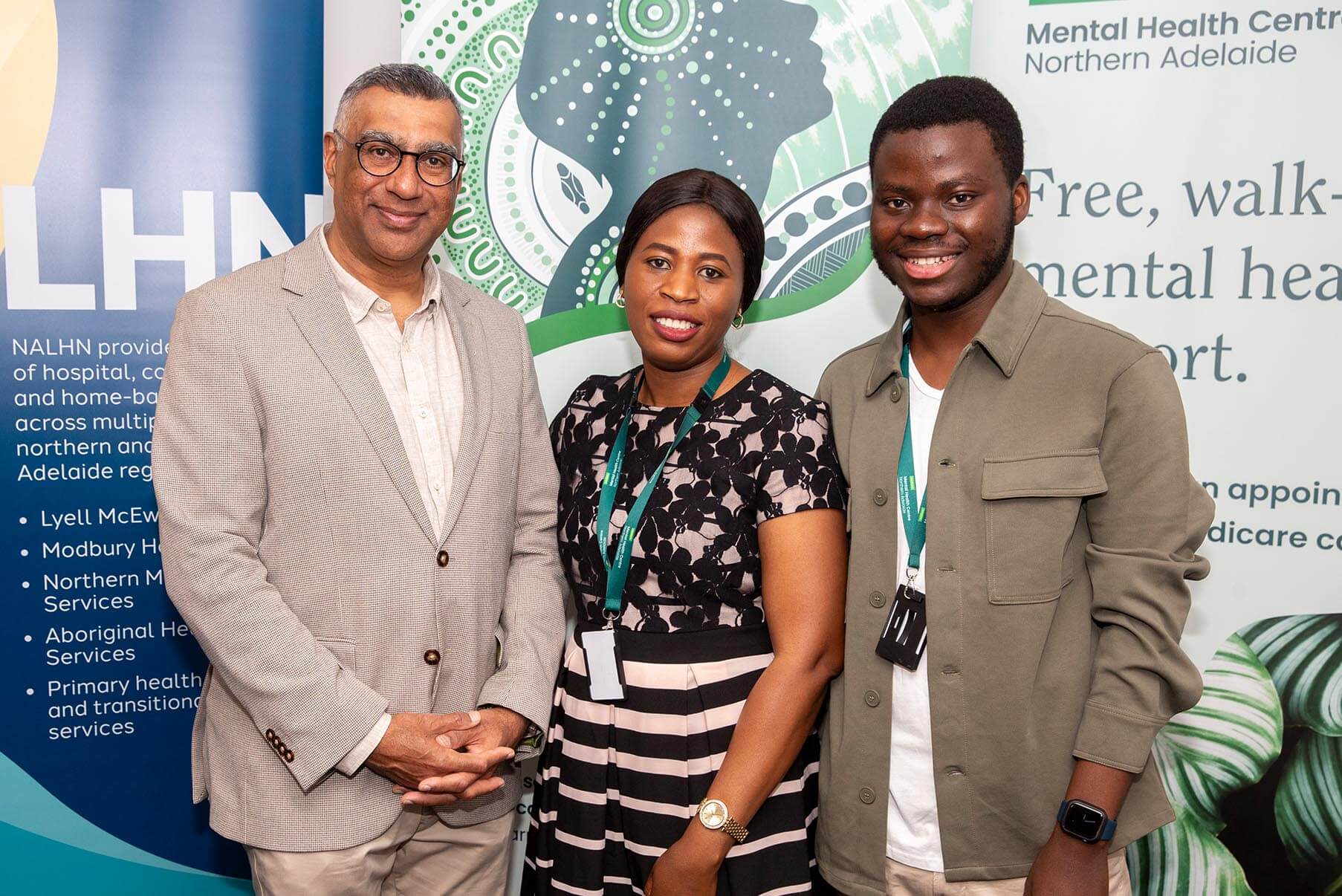 Three people stand together and smile in front of a banner at an event.