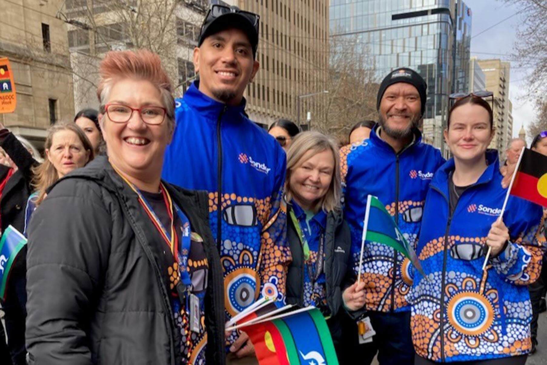 A woman with pink hair and others in blue jackets with orange patterns are smiling and holding flags.