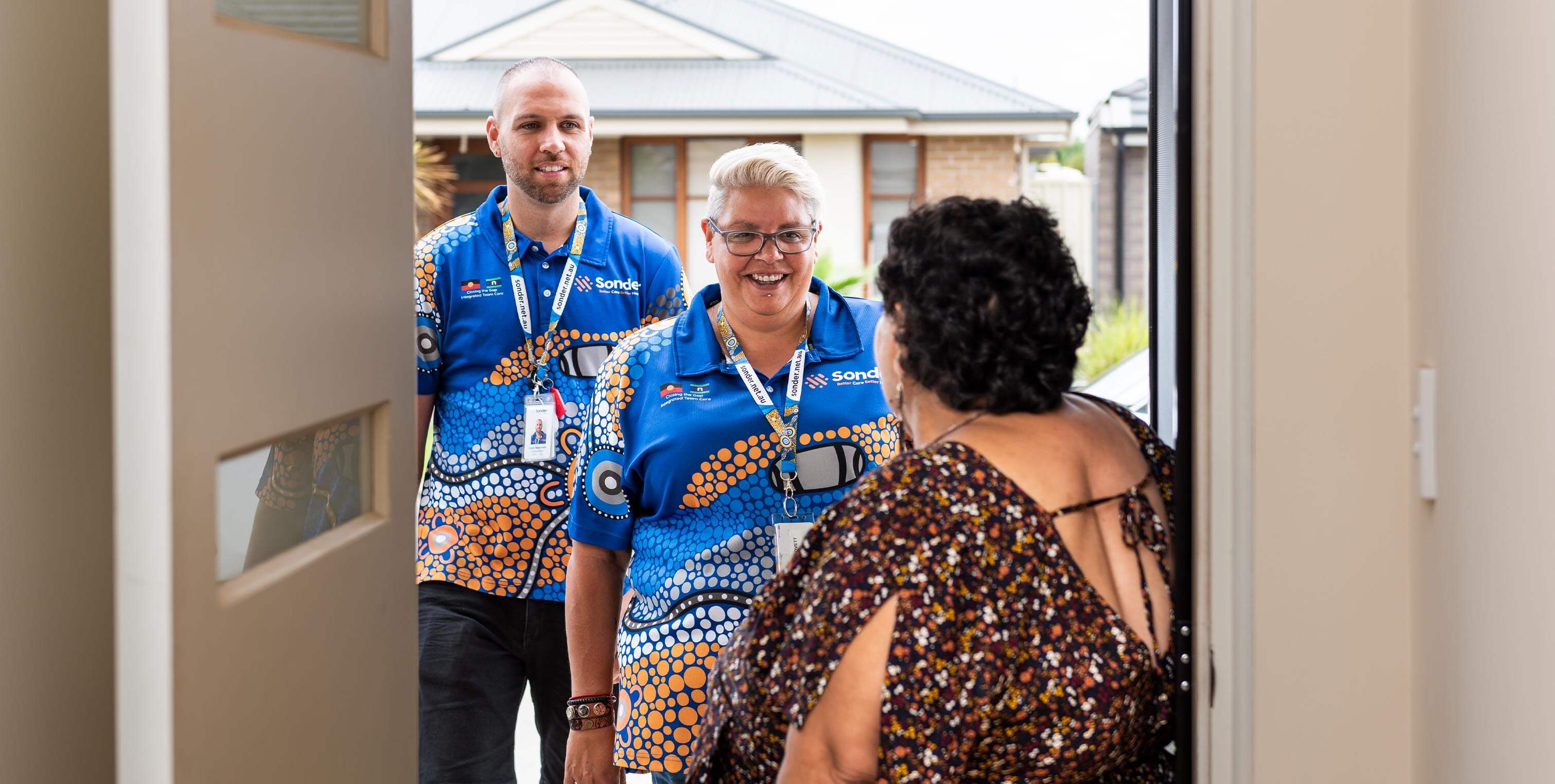 Two people wearing matching blue shirts with an Indigenous art pattern smile as they greet a person at a doorway.