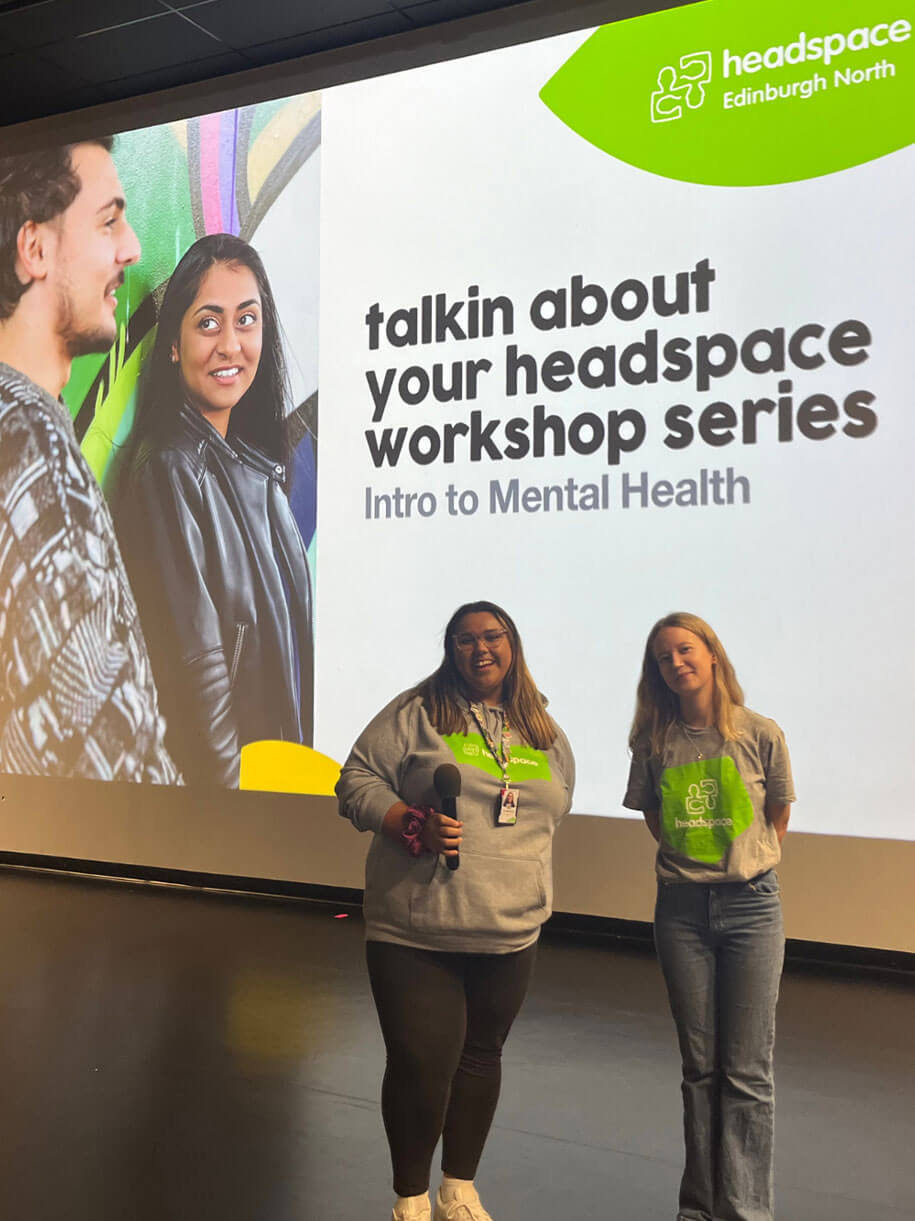 Two women, wearing shirts with the headspace logo, standing in front of a large screen at a workshop.